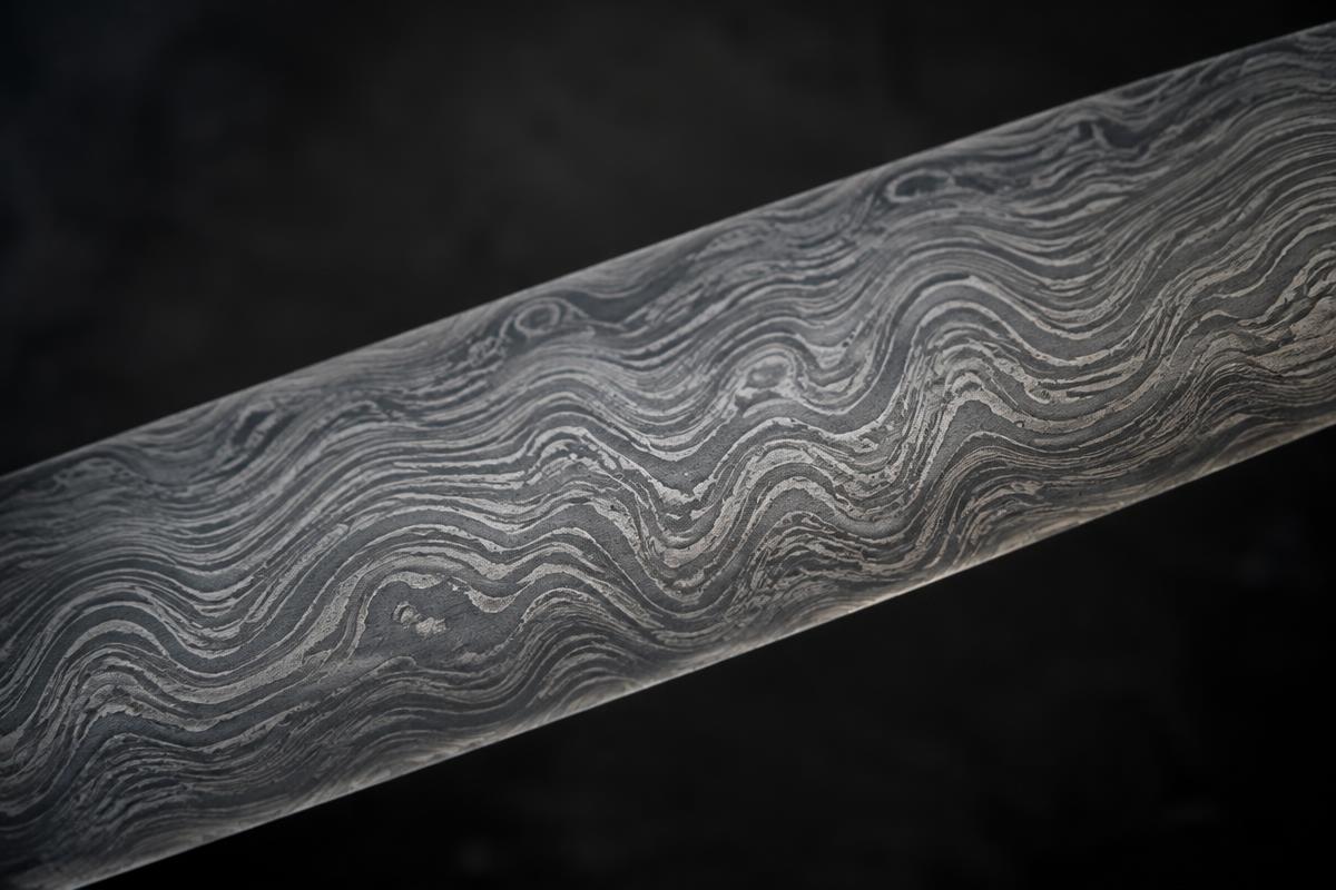 Close-up of a pattern-welded steel blade showing a flowing, layered wave pattern